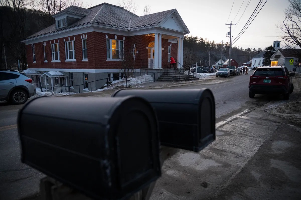 Two mailboxes in the foreground with a brick building on the opposite side of a snowy road. Cars are parked along the street, and a person is approaching the building's entrance.