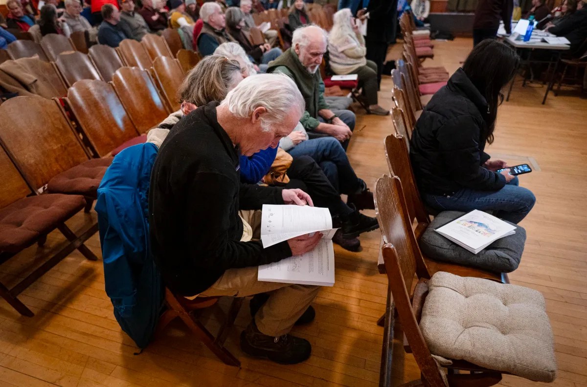 A group of people seated in a hall, some reading documents, others on phones. Wooden chairs and a wooden floor are visible.