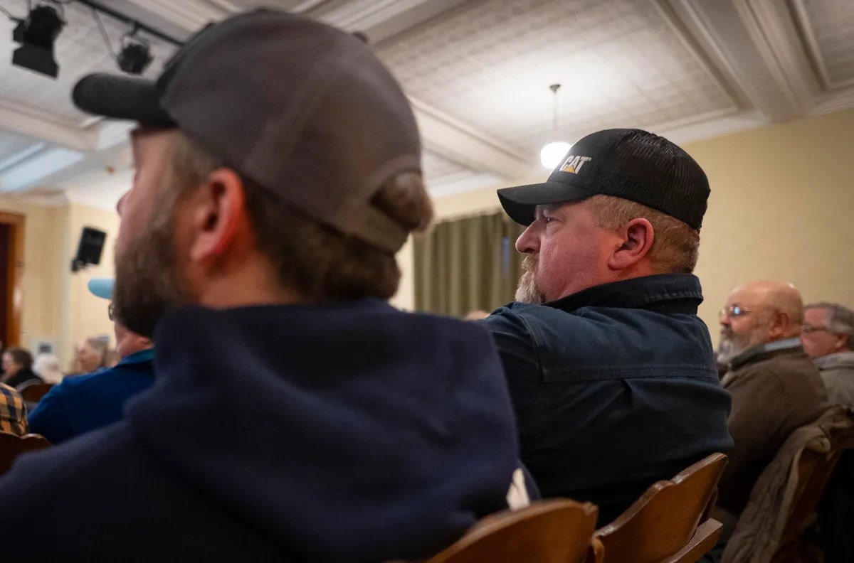 Audience seated in a room, focusing on an event or speaker off-camera. People wear hats and casual clothing.