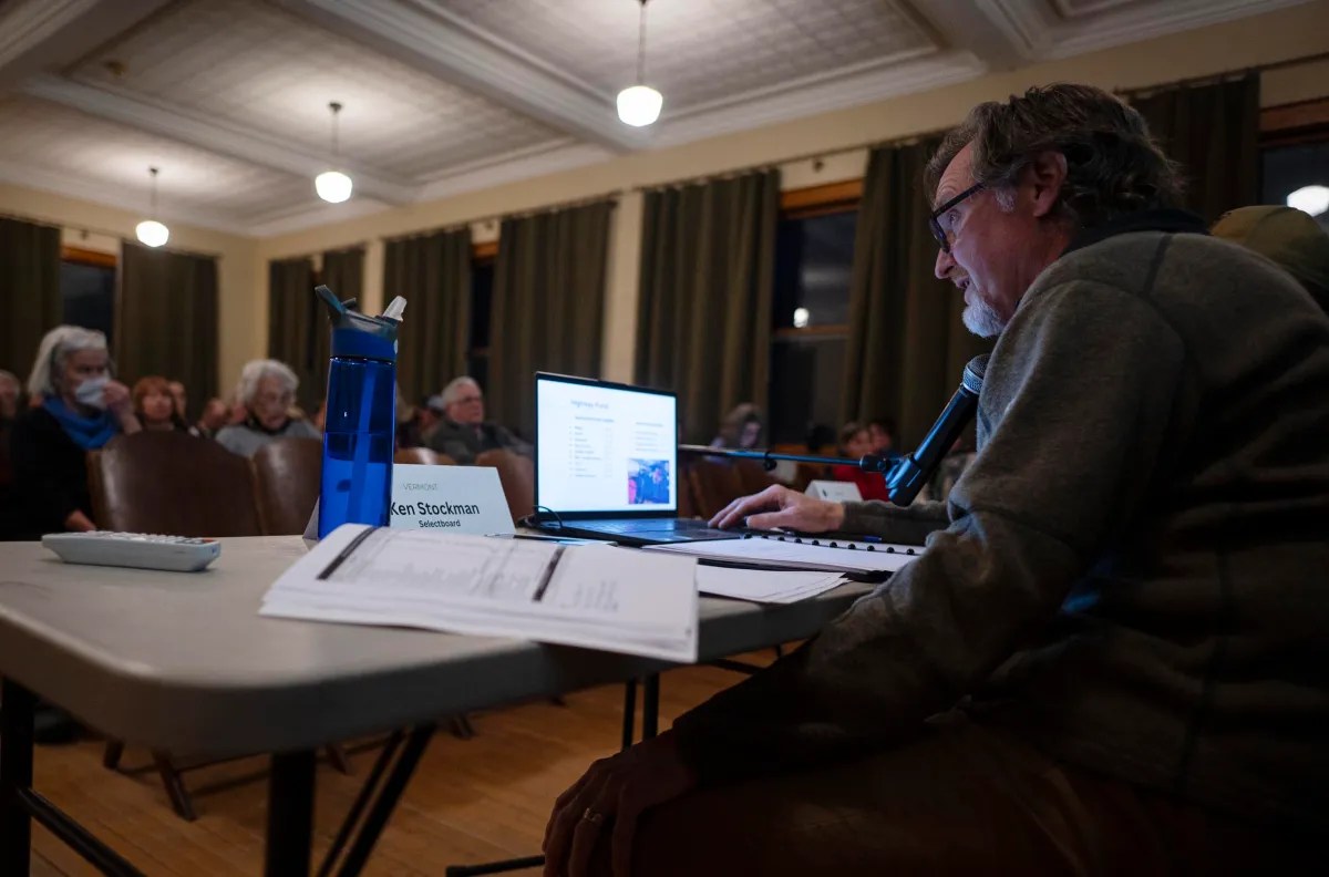 A person speaks into a microphone at a desk with a laptop in a dimly lit room, as attendees sit in the background listening.