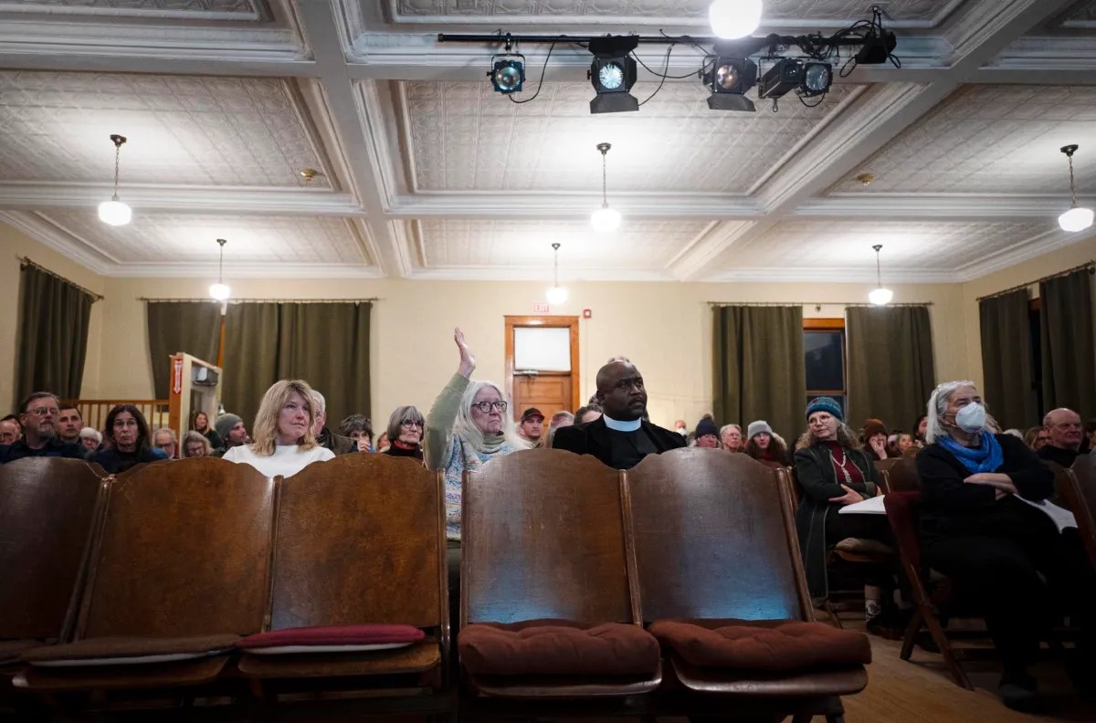 People sitting in a hall, some with raised hands, during a meeting. Chairs and light fixtures are visible.