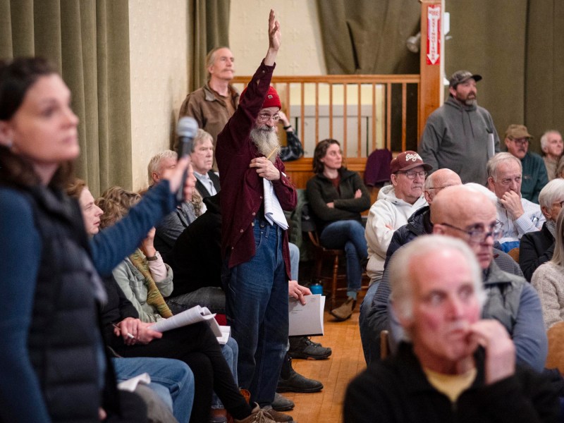 A man in a red hat raises his hand in a crowded meeting room while a woman holds a microphone. Other attendees sit and watch.