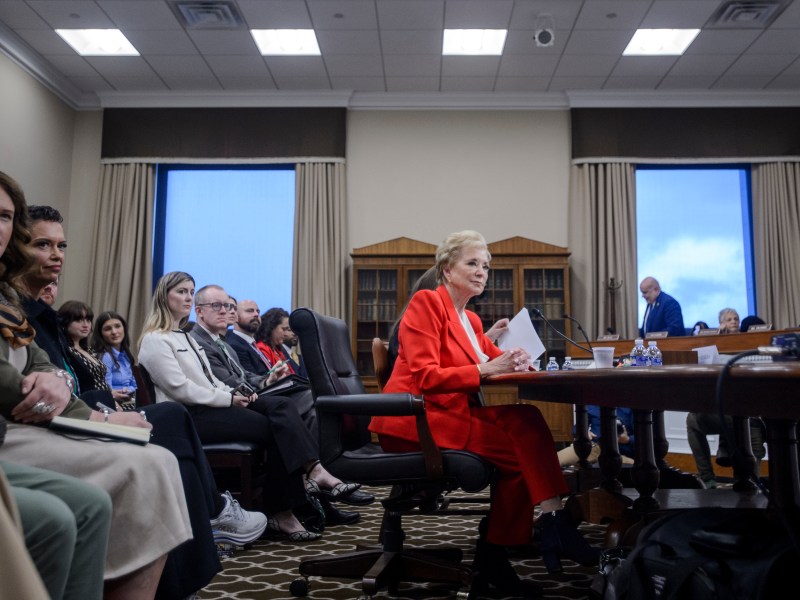 A woman in a red suit sits at a table speaking during a meeting or hearing, with several people seated and listening in a conference room.