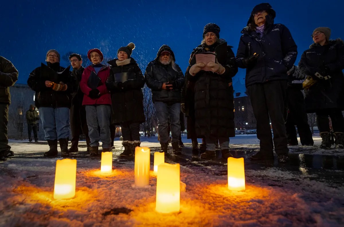 A group of people stand in a snowy area at dusk, holding candles. Several lit candles are placed on the ground in front of them.