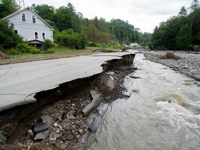 Damaged road with collapsed section beside a river after a flood, near a white house and surrounded by greenery.