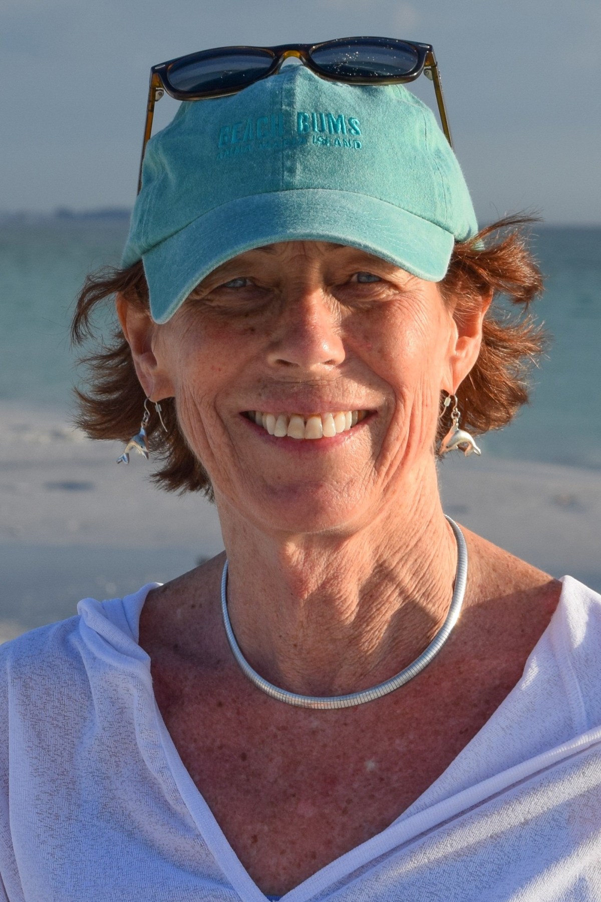A woman wearing a turquoise cap and sunglasses, white top, and silver necklace smiles on a sandy beach with the ocean in the background.