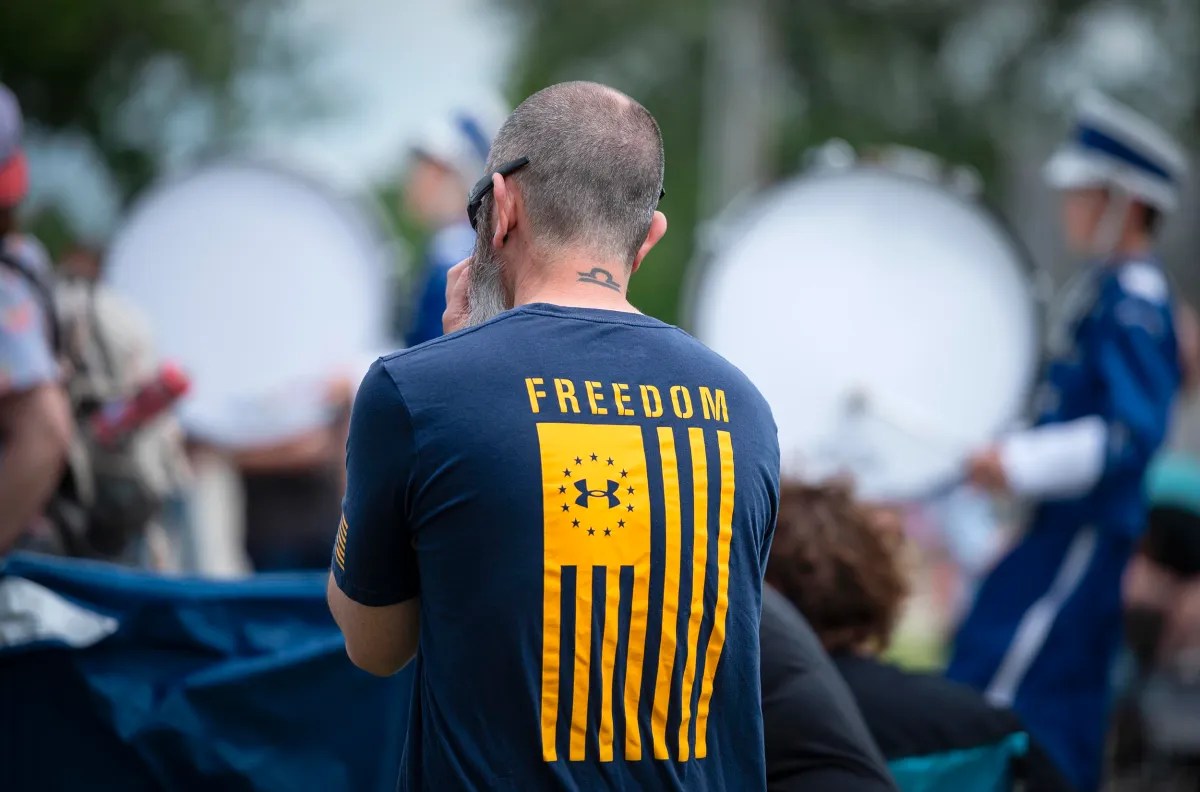 A man with a short haircut and a tattoo on the back of his neck stands outdoors, wearing a shirt with the word "FREEDOM" and an American flag design. Drumline musicians are in the background.