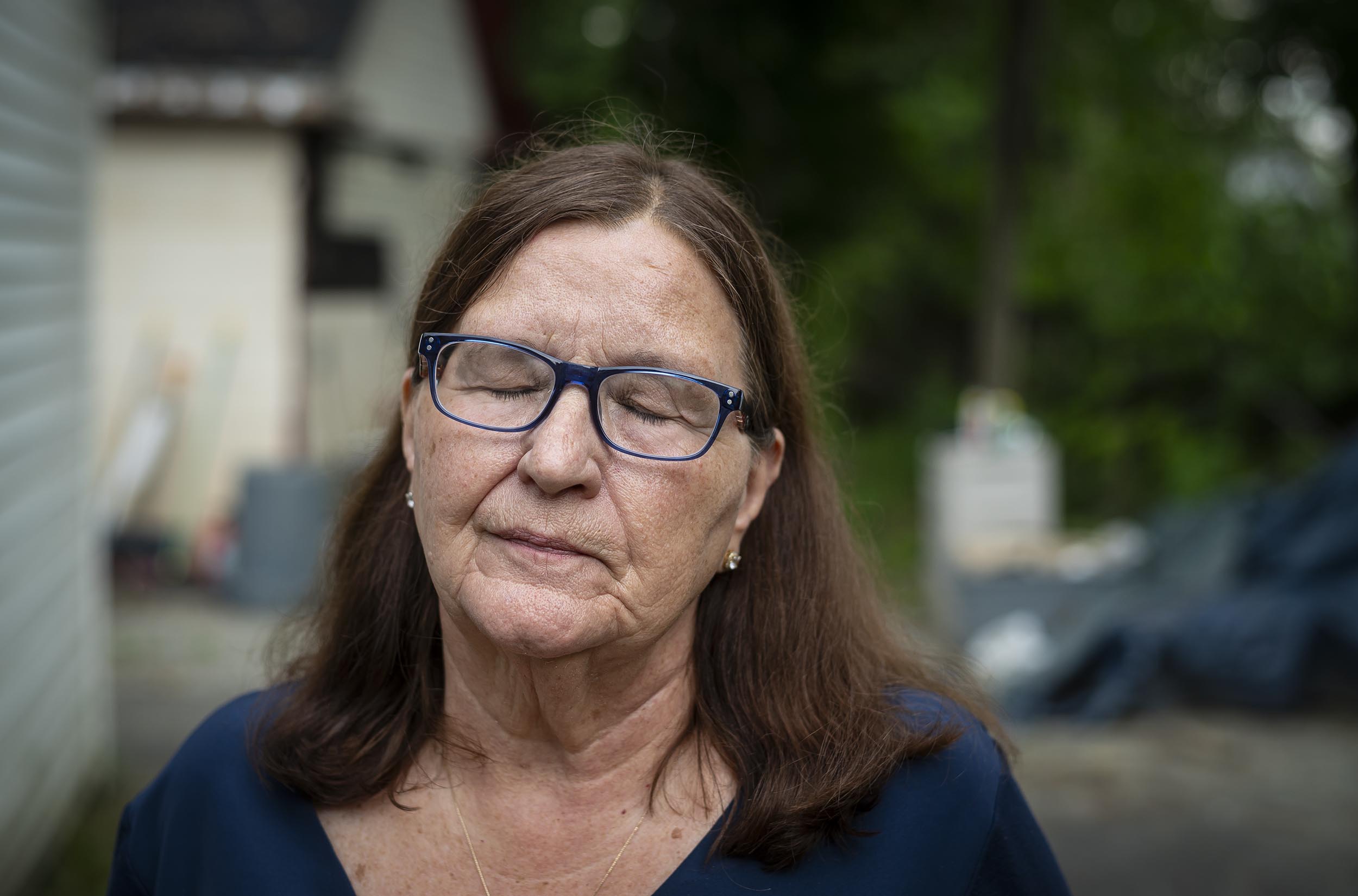 A woman with long brown hair and glasses is standing outdoors with her eyes closed. She is wearing a dark blue shirt. The background shows a building and greenery.