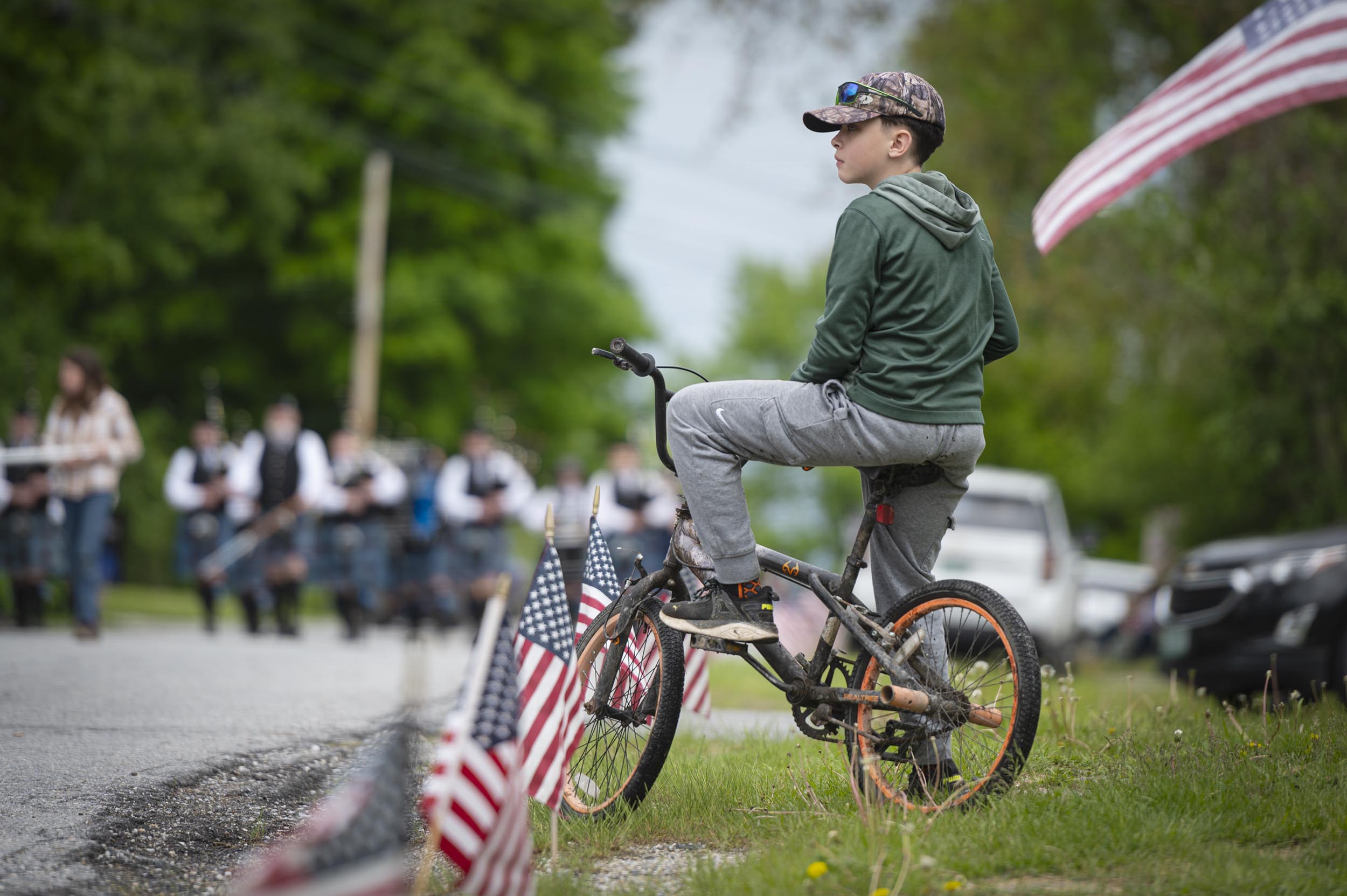 A boy in a green hoodie and cap sits on a bicycle, watching a parade with American flags lining the roadside.