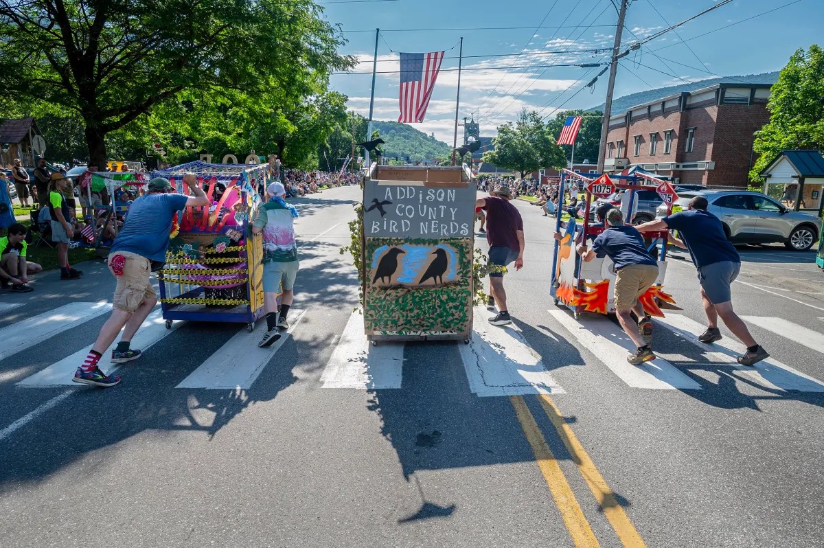 A group of people push ornately decorated carts labeled "Addison County Bird Nerds" across a crosswalk during a parade, with American flags and spectators in the background.