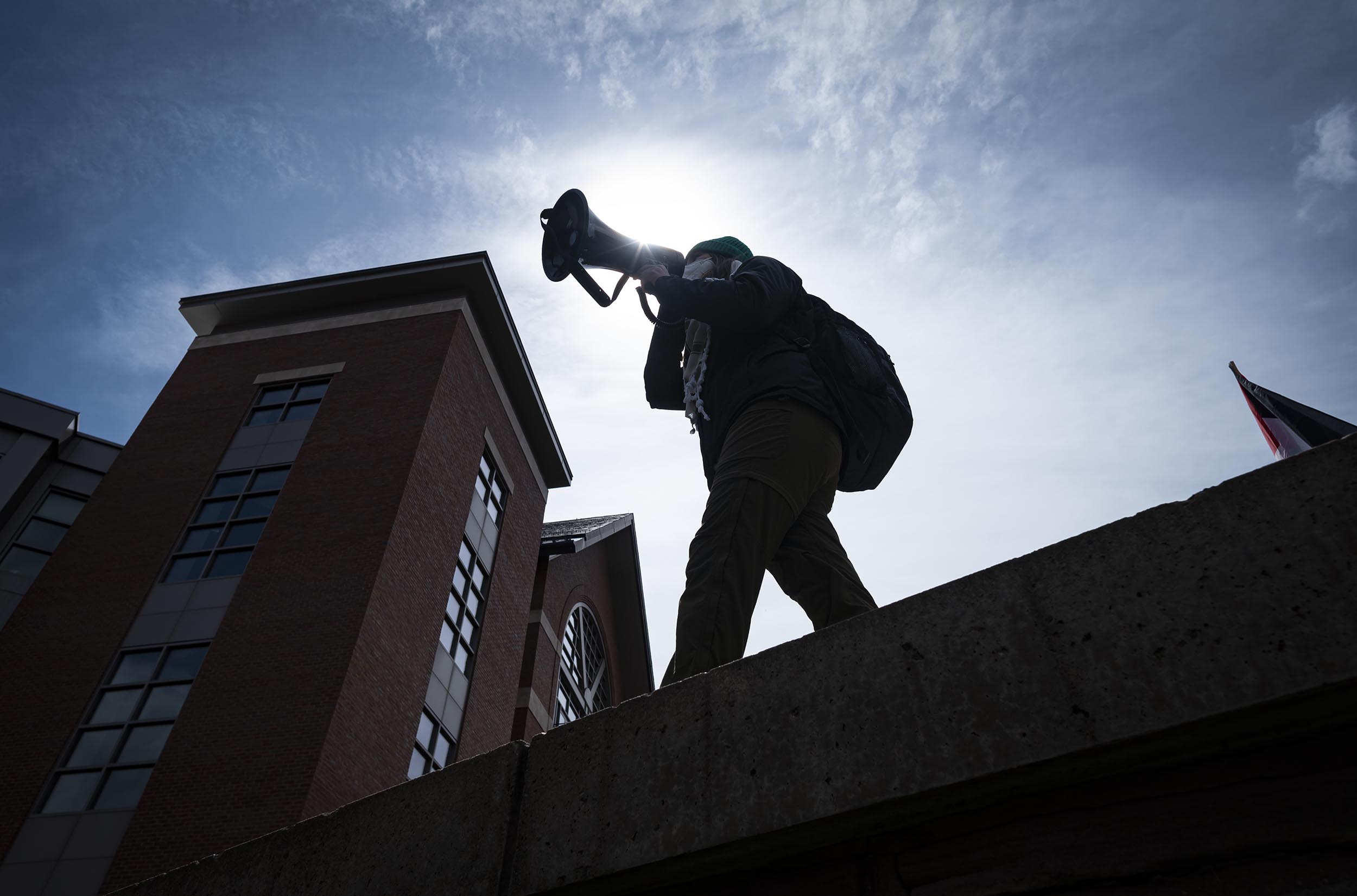 A protester wearing a helmet and backpack uses a megaphone against a sunlit sky, standing on an elevated surface with buildings in the background.