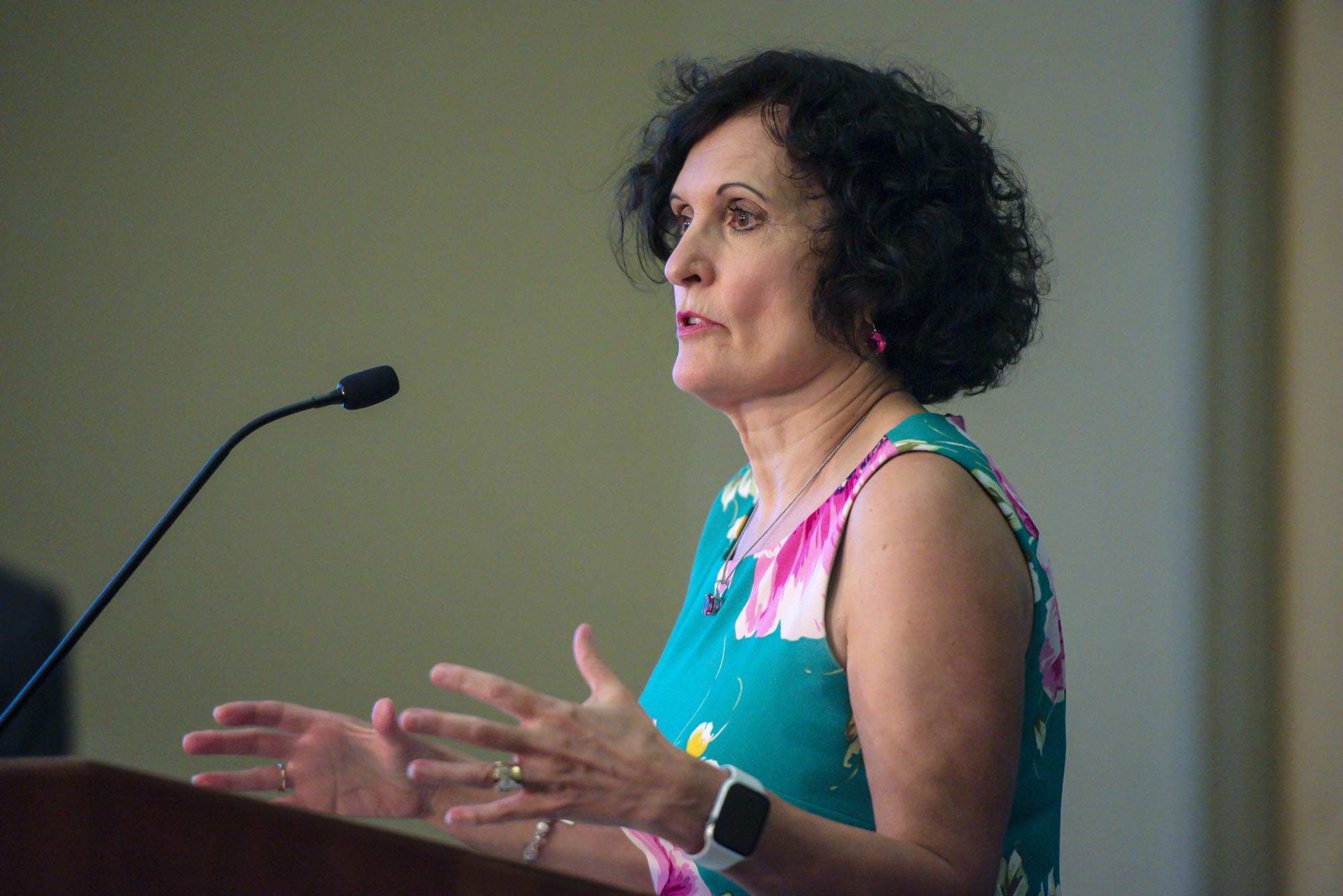 Person with short curly hair speaks at a podium, gesturing with hands. They wear a floral sleeveless top and a smartwatch.