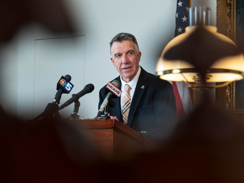 A man in a suit speaks at a podium with microphones in front of him, an American flag in the background, and a lamp in the foreground.