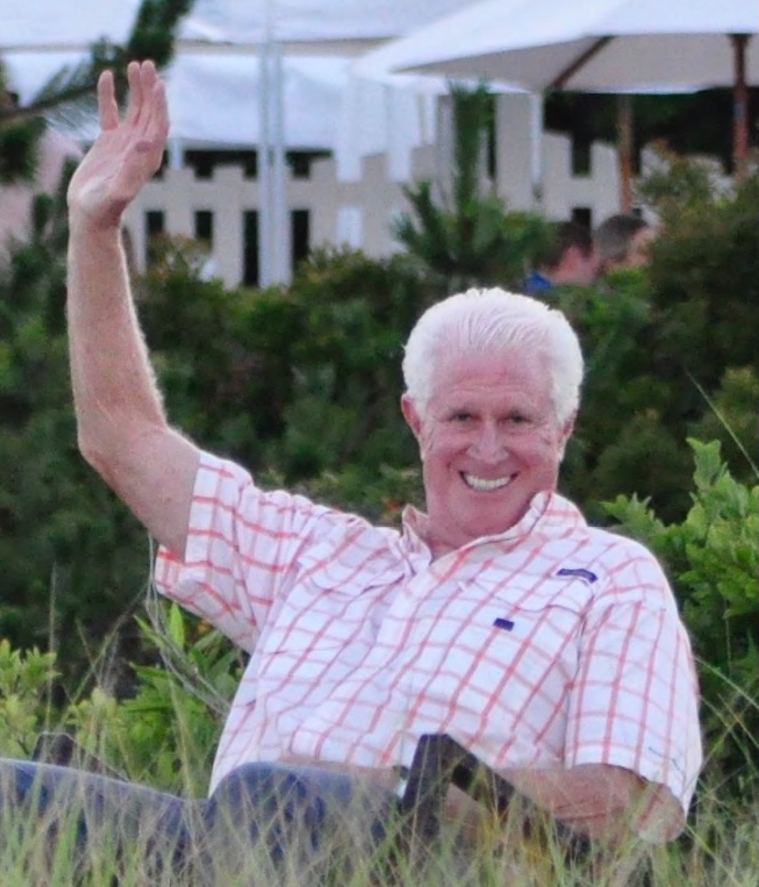 A man wearing a plaid shirt sits outdoors in a garden area, smiling and waving with his right hand.