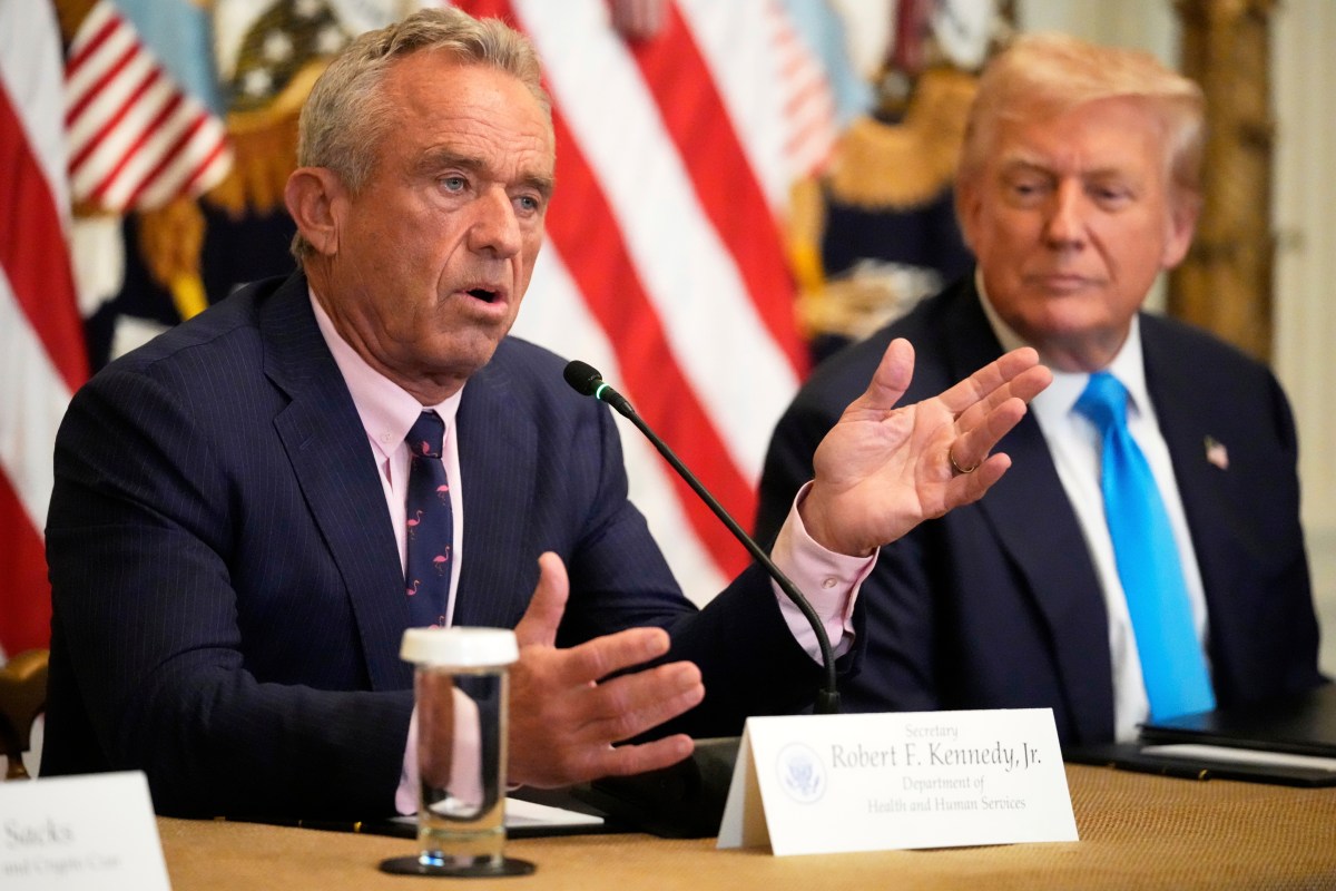 Two men in suits sit at a table with microphones; one is speaking while the other listens. American flags are visible in the background.