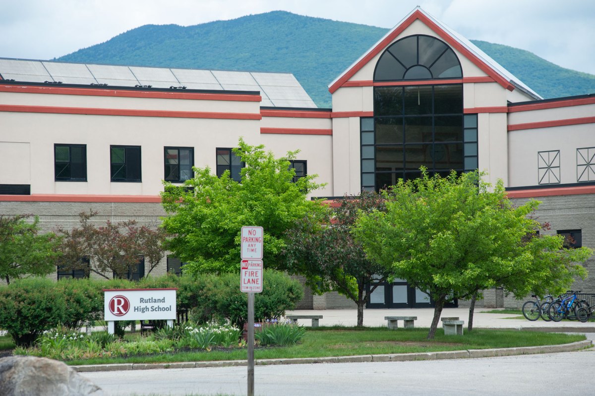 A beige and rust-colored building with large windows surrounded by trees and a lawn.