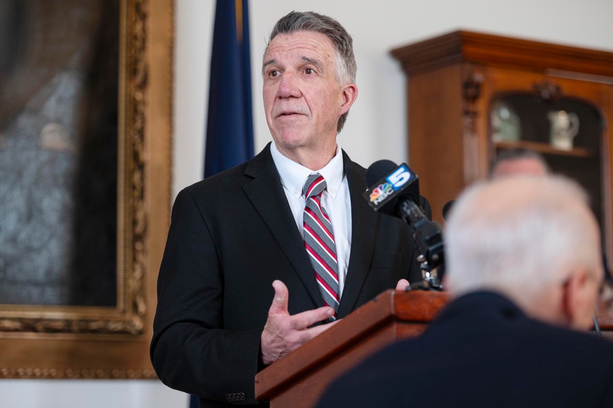 A man in a suit speaks at a podium with microphones, in an indoor setting with a flag and a wooden cabinet in the background.