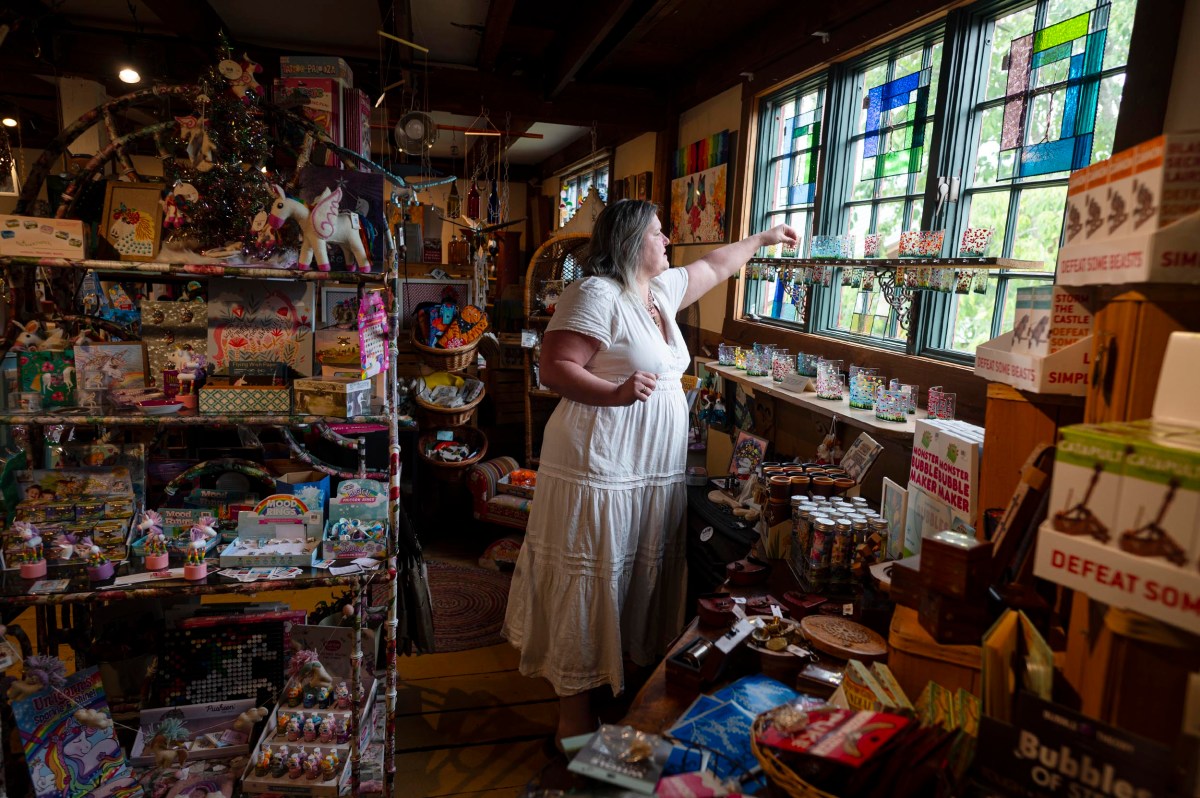 A woman in a white dress arranges items near a window inside a colorful, crowded gift shop filled with toys, trinkets, and stained glass decorations.
