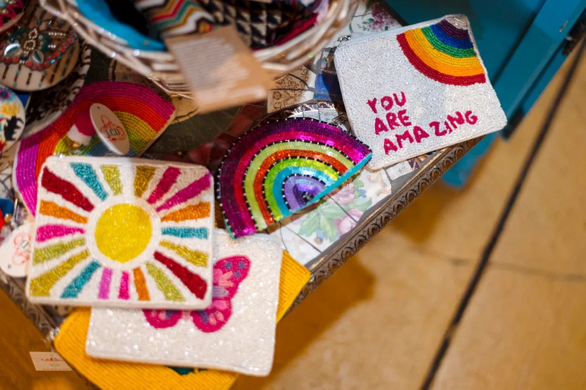 Several colorful, beaded pouches are displayed on a table, including ones with a sun, rainbow, butterfly, and the phrase "YOU ARE AMAZING.