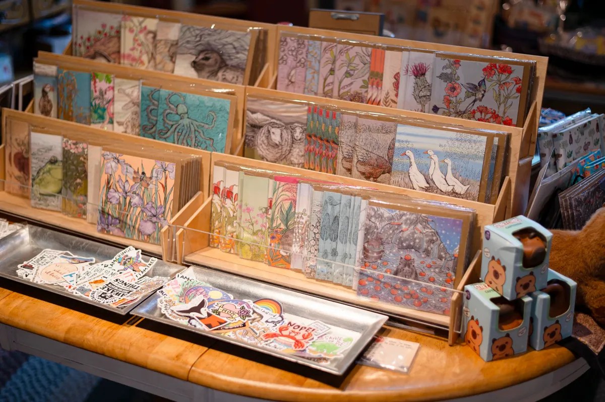 Display of illustrated greeting cards with nature themes and animal designs, arranged in wooden racks, alongside metal trays holding stickers and small boxed items on a wooden table.