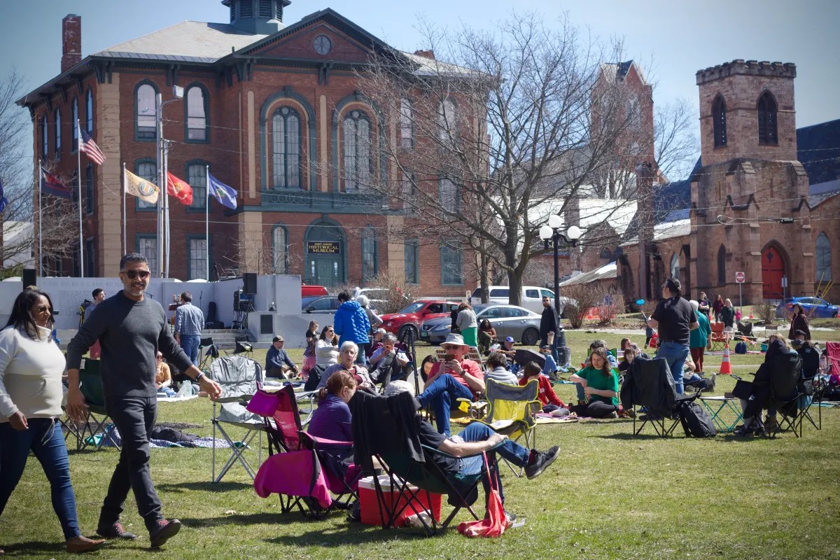 People gather on a grassy park in front of old brick and stone buildings.