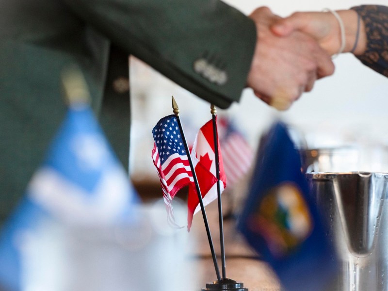 Two people shaking hands near small U.S. and Canada flags on a table, with blurred flags in the foreground.