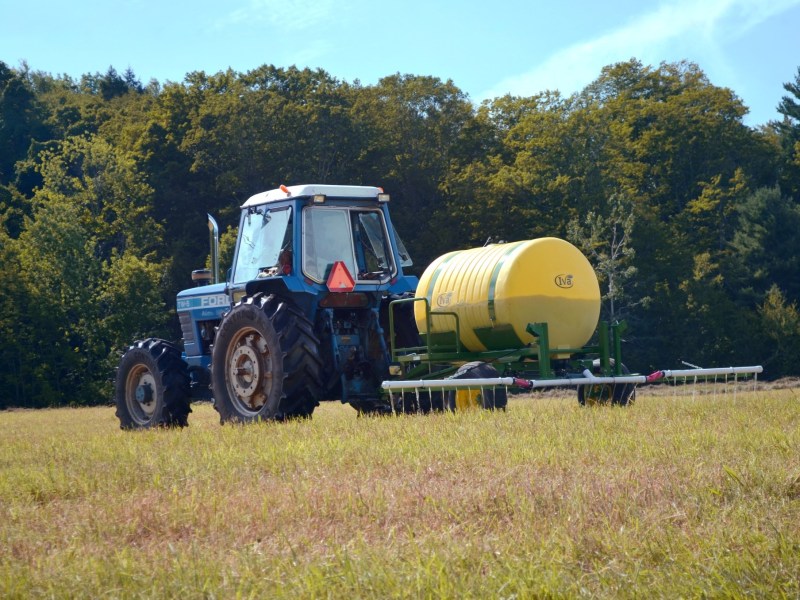 A blue tractor pulling a yellow tank sprayer across a grassy field with trees in the background under a clear sky.