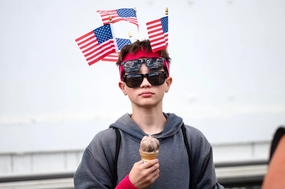 A person wearing sunglasses, a red headband, and several small American flags on their head holds an ice cream cone.