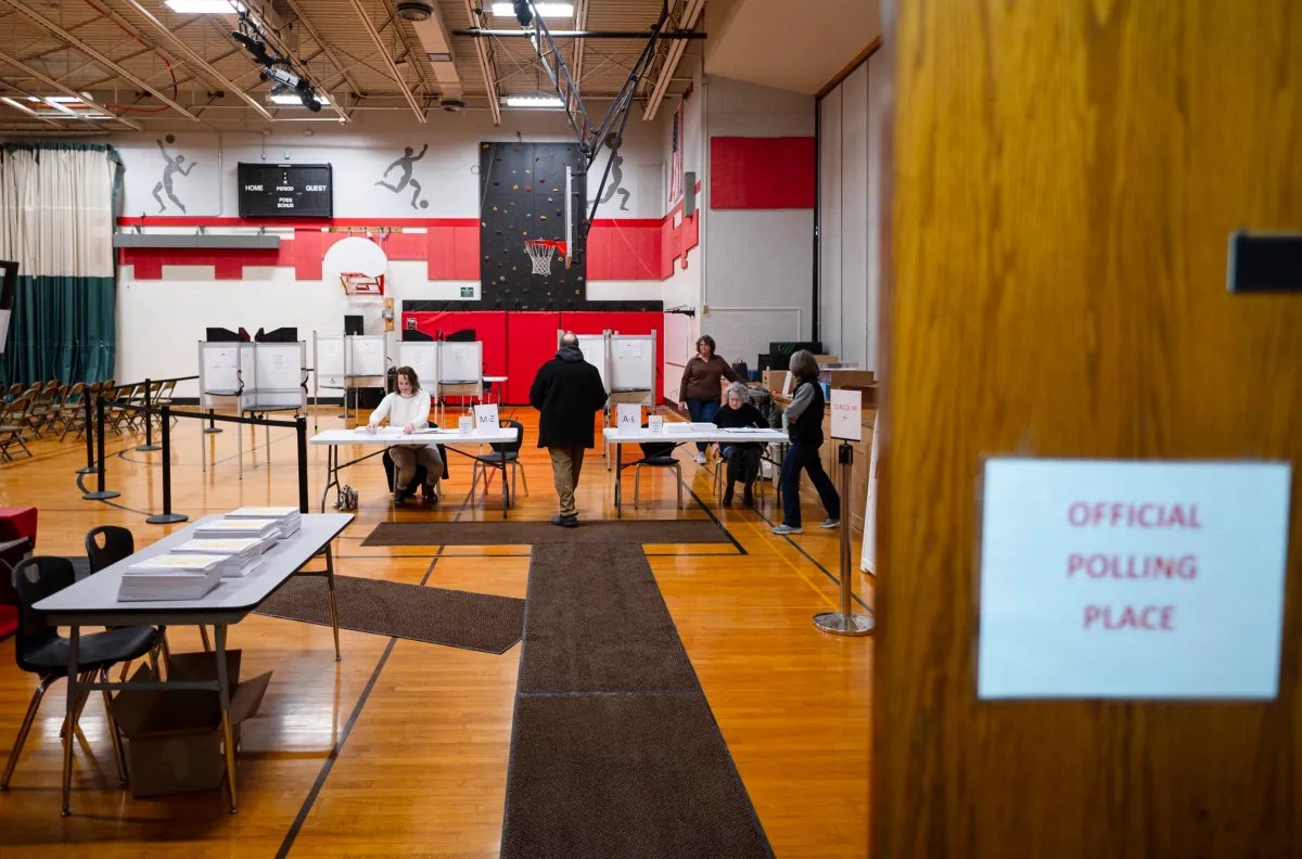 A man at a polling station inside a gymnasium, approaching workers seated at tables with voting booths in the background. A sign on the door reads "Official Polling Place.