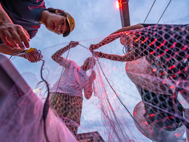 Three people repairing a fishing net under red light against a blue sky.