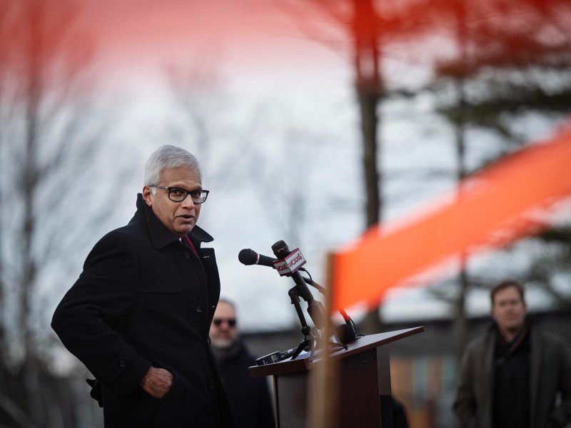 A person in a black coat speaks at a podium with microphones outdoors on a cloudy day. An orange ribbon is visible in the foreground.