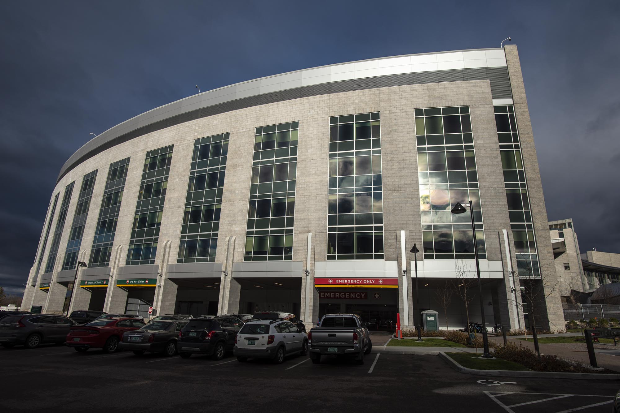 Exterior view of a multi-story hospital building with cars parked in the lot and an emergency entrance visible.