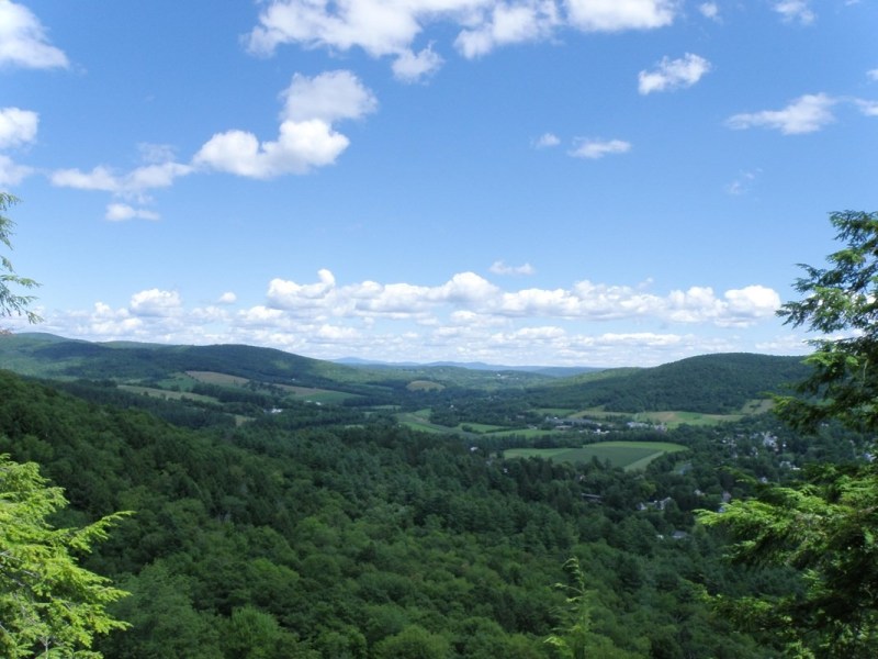 A panoramic view of rolling green hills and forests under a blue sky with scattered white clouds.