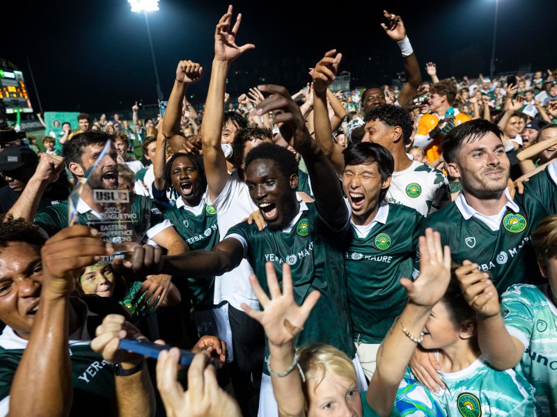 A soccer team in green uniforms celebrates on the field at night, holding a trophy and surrounded by cheering fans.