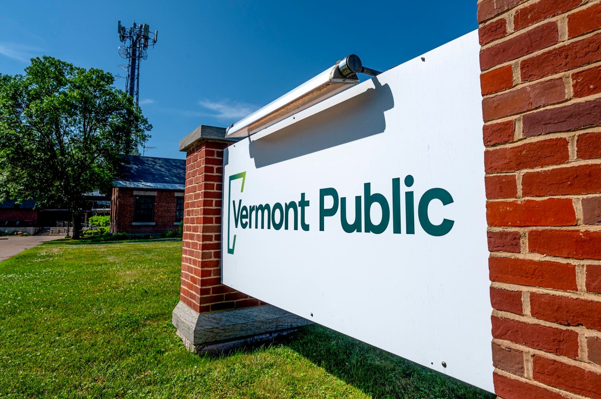 A white sign reading "Vermont Public" is mounted between two brick pillars outside, with a grassy area and a communication tower in the background.