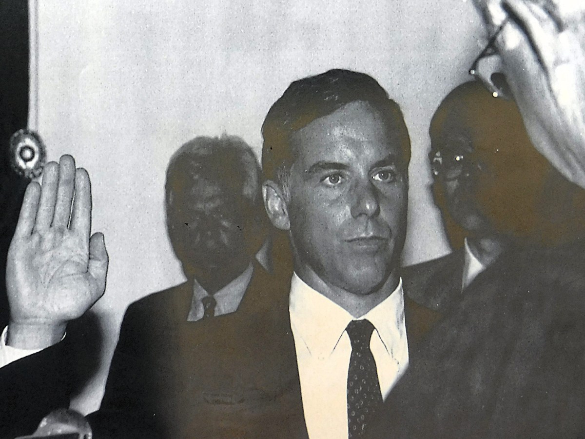A man in a suit raises his right hand while being sworn in, with two other men standing in the background. The image is in black and white.
