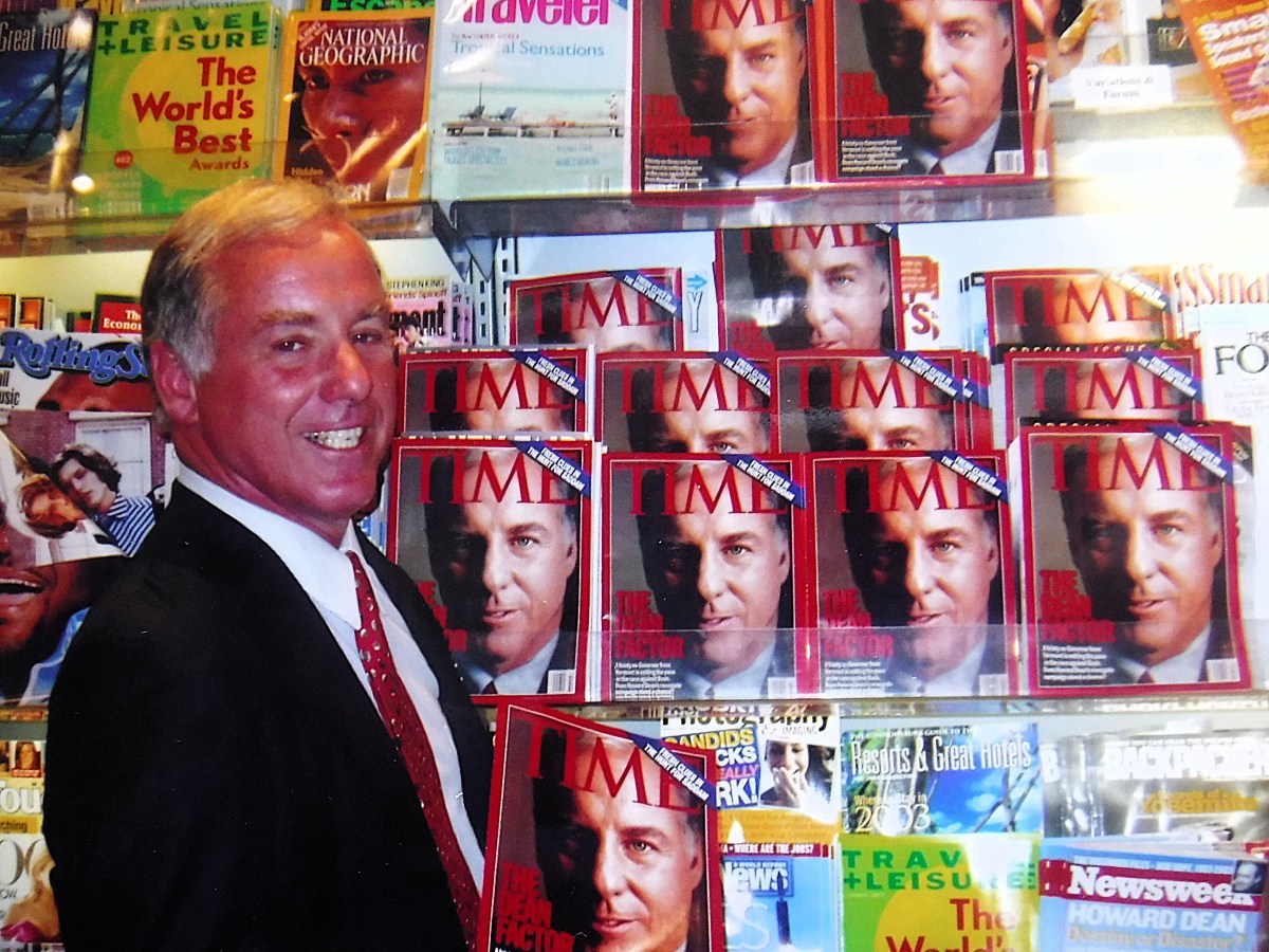 A man in a suit smiles while standing in front of a magazine rack displaying multiple copies of a TIME magazine issue featuring his face on the cover.