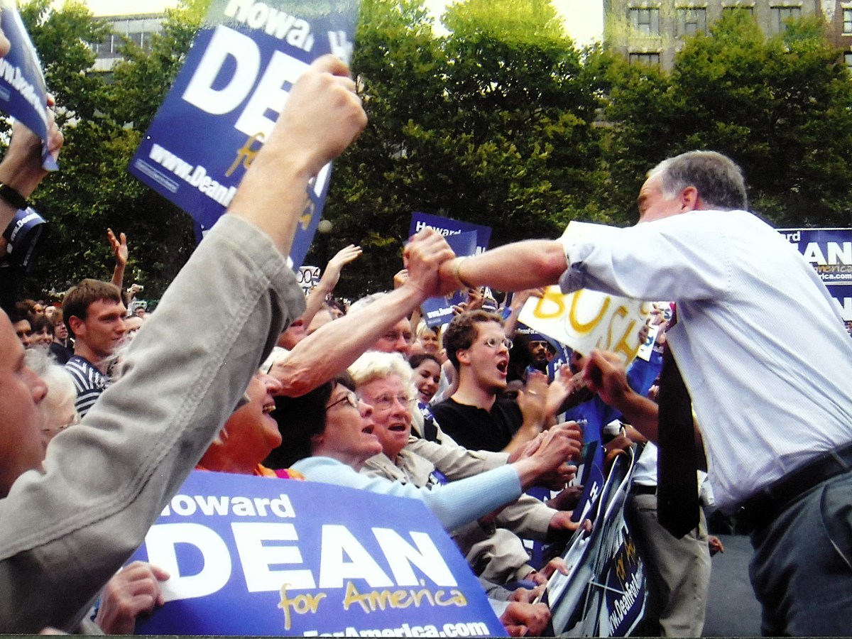Howard Dean greets and shakes hands with enthusiastic supporters holding "Howard Dean for America" signs at an outdoor campaign event.