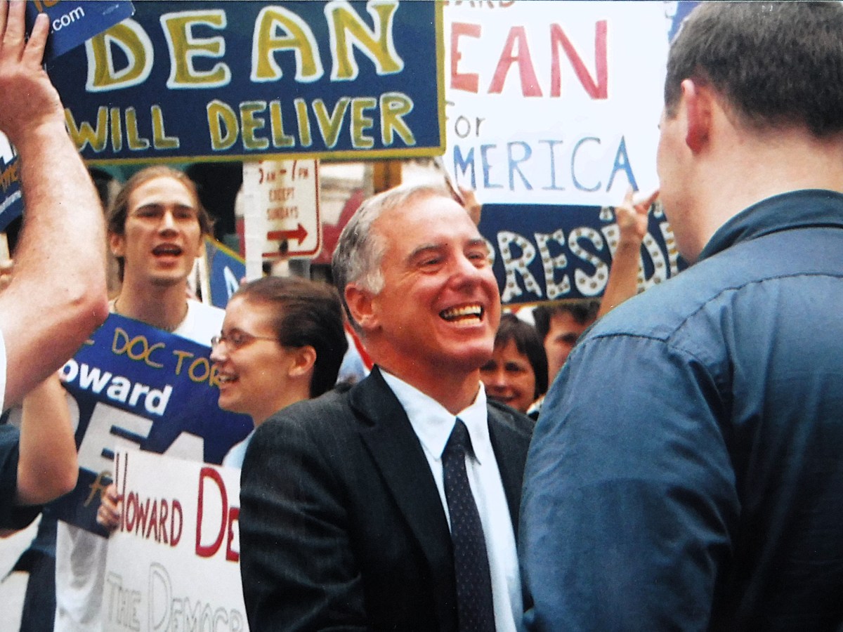 A man in a suit smiles and shakes hands with another person among a crowd holding campaign signs and banners.