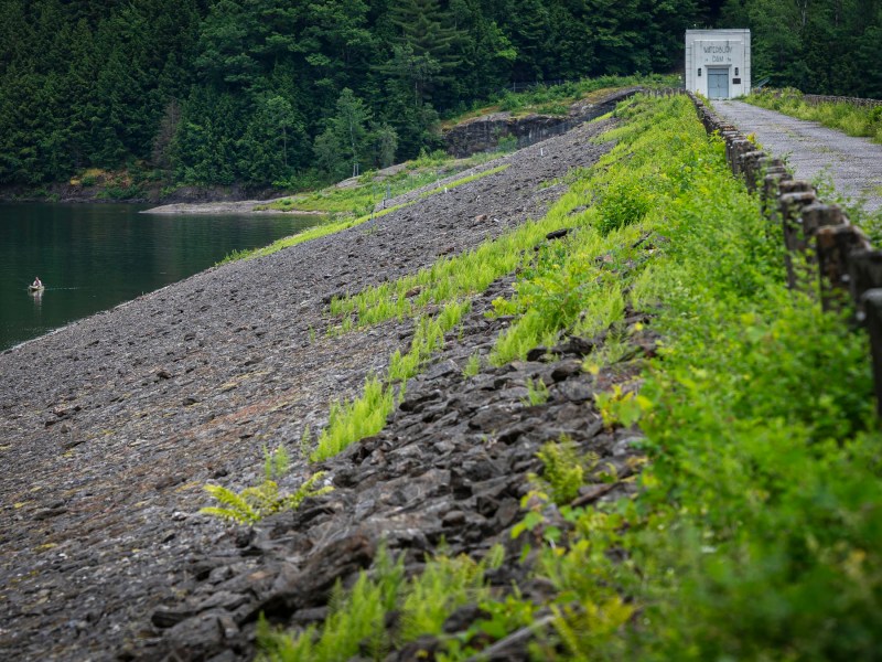 A concrete structure stands at the end of a gravel path atop a sloped dam covered with rock and patches of green vegetation, adjacent to a calm lake with a small boat near the shore.