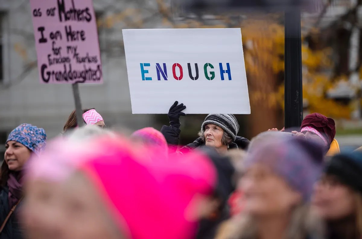 A group of people at a protest, with a prominent sign reading "ENOUGH" held by a person wearing a hat.
