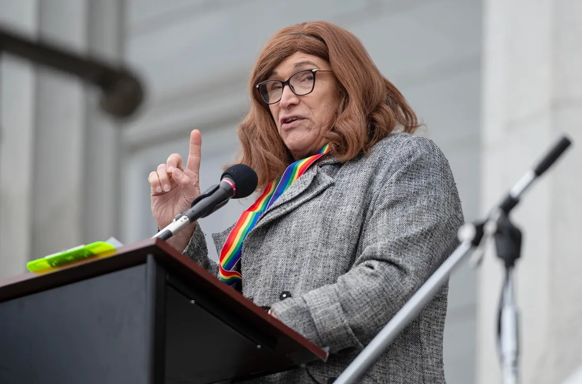 Person with long brown hair speaking at a podium, wearing glasses, a gray coat, and a colorful scarf.