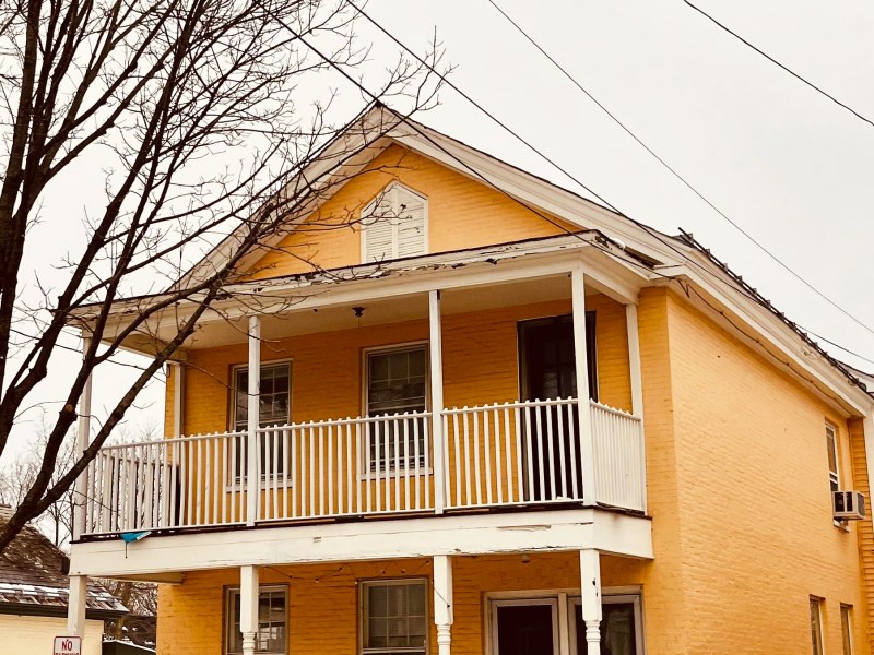 A two-story yellow house with white trim and a porch, featuring a front balcony and leafless trees nearby.