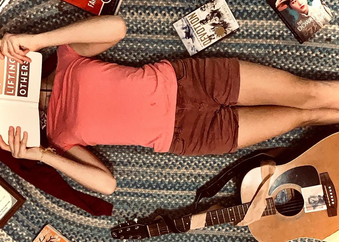 Person lying on a blue patterned rug, reading a book, surrounded by scattered books and an acoustic guitar.