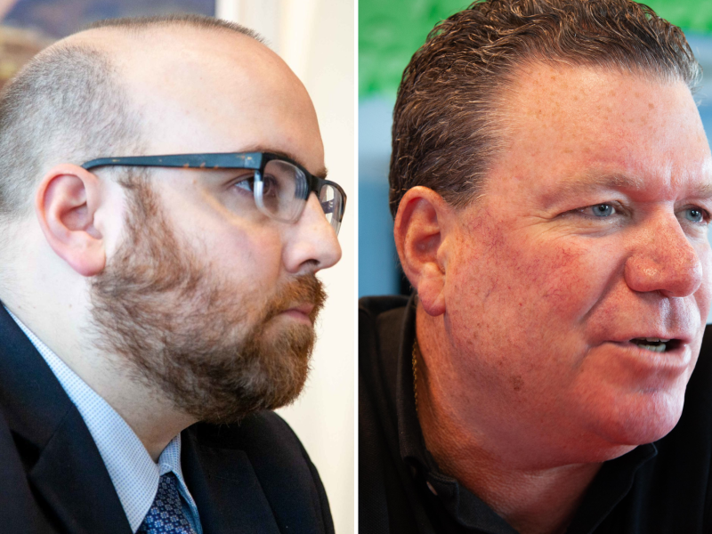 Two men in profile view. The man on the left wears glasses and a suit, and the man on the right has a casual shirt. Both have facial hair and are indoors.
