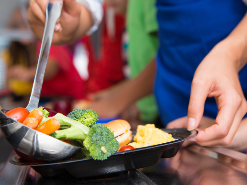Person serving vegetables, a sandwich, and scrambled eggs onto a tray in a cafeteria setting.